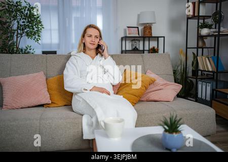 Frau, die auf dem Smartphone im Bademantel spricht und sich zu Hause auf der Couch entspannt Stockfoto