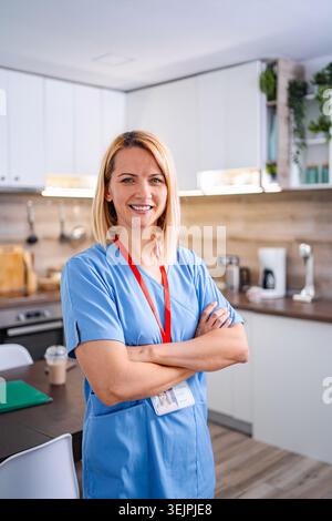 Selbstbewusste Ärztin in blauer Uniform, die mit überkreuzten Armen in der Küche steht Stockfoto