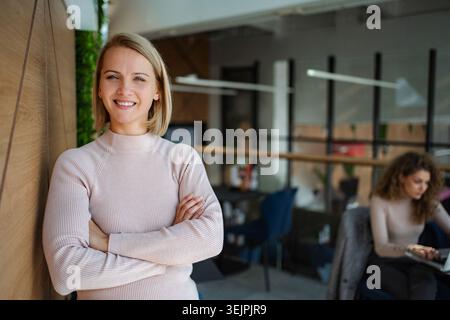 Selbstbewusste Geschäftsfrau lächelt mit überkreuzten Armen im modernen Büro Stockfoto