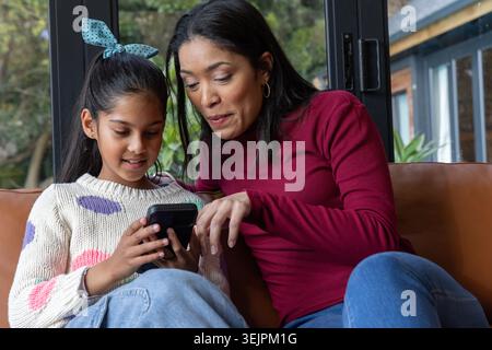 Asiatische Mutter und Kind sitzen im Wohnzimmer auf brauner Ledercouch und halten das Smartphone zusammen. Familie, Eltern, Mädchen, gemütlich, ich Stockfoto