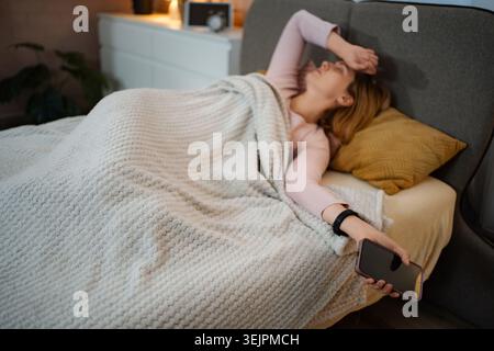Müde Frau, die im Bett liegt und ein Smartphone hält Stockfoto