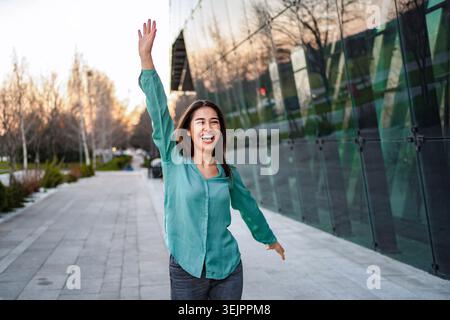 Junge Geschäftsfrau winkt und lacht vor einem modernen Bürogebäude Stockfoto