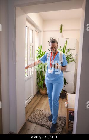 Lächelnde Ärztin, die blaue Peelings trägt, verlässt das Haus zur Arbeit und überprüft ihr Telefon Stockfoto