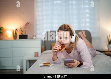 Frau isst Kuchen im Bett, während sie Musik hört Stockfoto