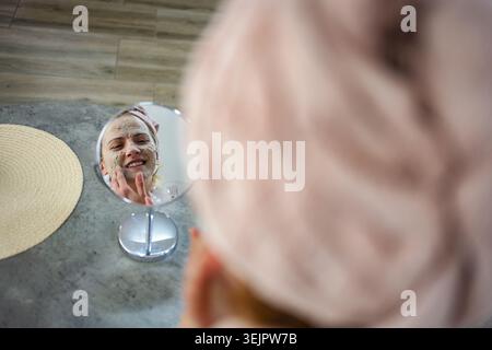 Frau, die eine Gesichtsmaske aufträgt und in den Spiegel schaut Stockfoto