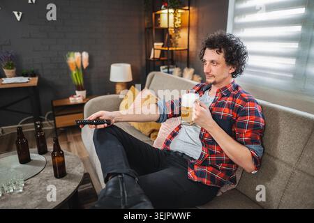 Junger Mann, der sich auf dem Sofa entspannt, Bier trinkt und fernsieht Stockfoto