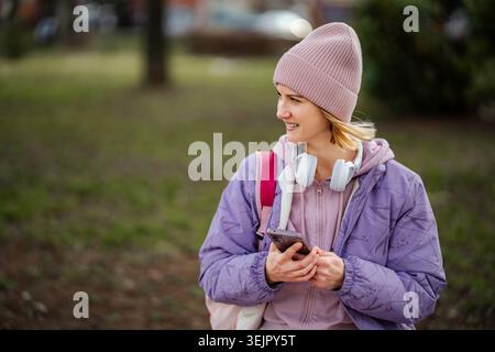 Junge Frau lächelt mit Smartphone und trägt Beanie-Kopfhörer Stockfoto