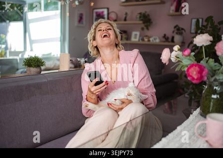 Eine lachende Frau hält Telefon und Katze auf der Couch Stockfoto