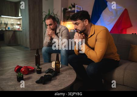 Freunde sehen sich ein aufregendes Match an, während sie Getränke und Snacks in einem gemütlichen Wohnzimmer genießen, das mit einer französischen Flagge geschmückt ist Stockfoto