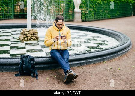 Junger attraktiver bärtiger Mann in lässiger Kleidung, der neben Steinbrunnen sitzt und auf den Bildschirm seines Smartphone-Watts konzentriert wird Stockfoto