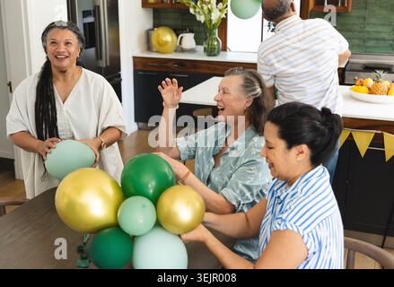 Vielfältige reife Gruppe, die Ballons aus grünem Gold in Teal in der Küche zuhause in lässiger Kleidung zubereiten. Familie, Einrichtung, Party, Celebrati Stockfoto