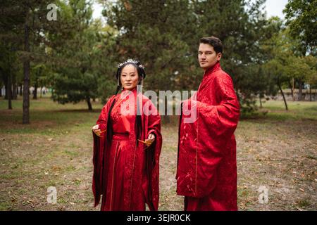 Interkulturelles Paar feiert Hochzeit in traditionellen chinesischen Gewändern im Freien Stockfoto