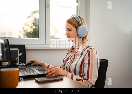Frau, die ein Headset trägt und vom Home Office aus am Laptop arbeitet Stockfoto