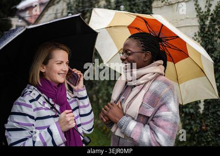 Verschiedene Freundinnen unterhalten sich mit Regenschirmen und lachen draußen Stockfoto