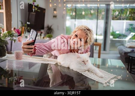 Eine lächelnde Frau nimmt Selfie mit einer Katze zu Hause Stockfoto
