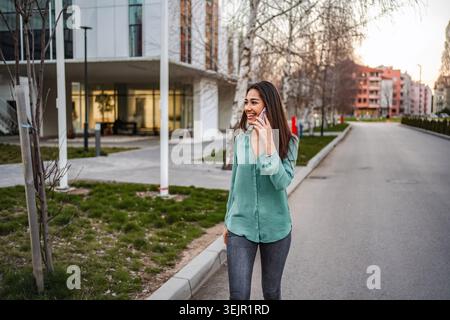 Junge Geschäftsfrau, die die Straße entlang läuft, während sie mit dem Smartphone spricht Stockfoto