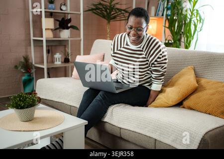 Lächelnde schwarze Frau, die sich zu Hause auf dem Sofa mit dem Laptop entspannt Stockfoto