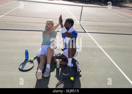 Diverse Paare in Sportbekleidung sitzen auf dem Tennisplatz über das Netz und geben High Five mit Schlägern, Bälle. Duo, draußen, sonnig, Schläger Stockfoto
