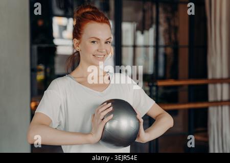 Schöne Ingwerfrau in Sportkleidung lächelt vor der Kamera und hält den silbernen kleinen Fitball in den Händen Stockfoto