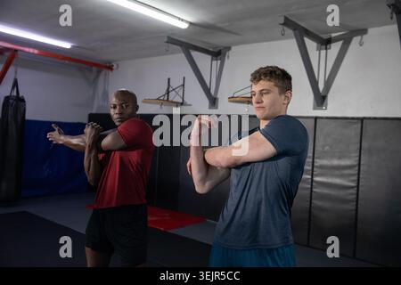 Verschiedene männliche Trainingspartner dehnen die Arme in Dojo auf Matten neben Boxsack in Sportbekleidung. Fitnessstudio, Fitness, Duo, Schulter, Krieg Stockfoto