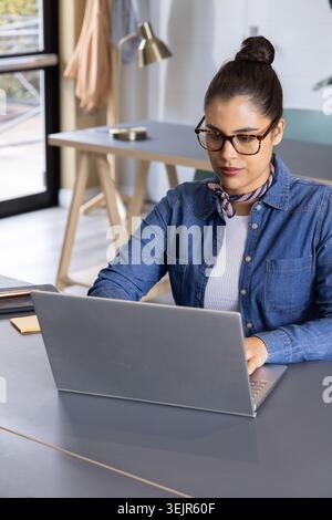 Frau, die im Home Office an schlanken silbernen Laptops arbeitet, Denimhemd und Schildpatt-Brille trägt. Arbeitsbereich, Schreibtisch, Lampe, Tagesleuchte Stockfoto