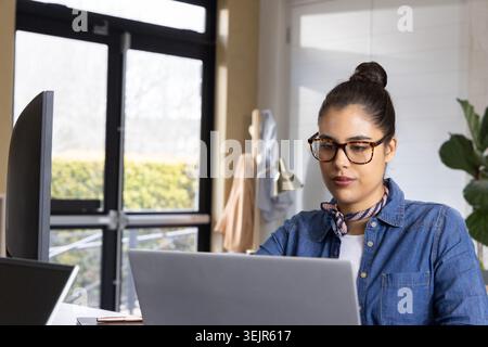 Frau, die am Laptop am Schreibtisch in einem hellen Büro am Fenster arbeitet, mit Denim-Hemd, Kopierraum. Workstation, Monitor, Computer, Glas Stockfoto