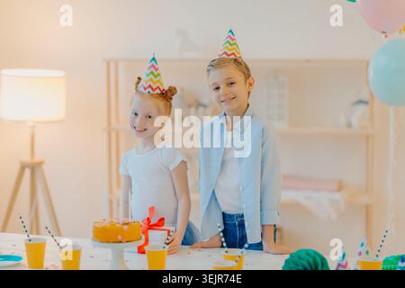 Zwei Kinder tragen Partyhüte, die zu Hause neben der Geburtstagskuche stehen und einen Geschenktisch tragen Stockfoto