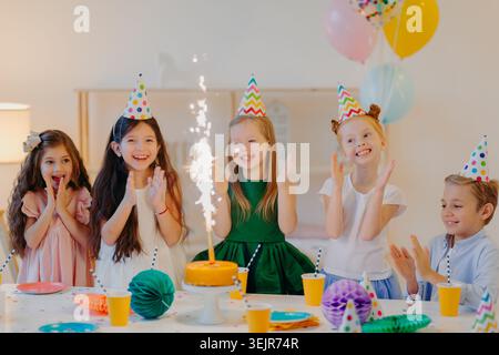 Gruppe von Kindern mit Partyhüten, die zu Hause Geburtstag mit Kuchen und Sparkler feiern Stockfoto