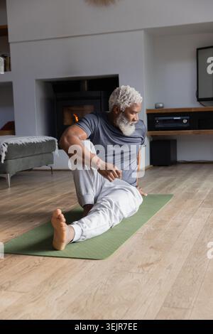 Senior Afroamerikaner, der sitzende Twist auf grüner Matte im Wohnzimmer mit Jogginghosen macht. Yoga, Dehnen, Kamin, h Stockfoto