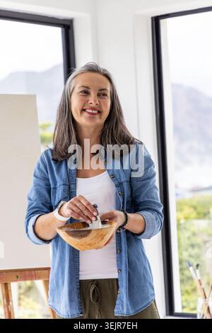 Frau mittleren Alters in Denim-Hemd und Olive-Hose in der Studioecke, die Farbe mit einem Palettenmesser mischt. Maler, Leinwand, Staffelei, Pigm Stockfoto