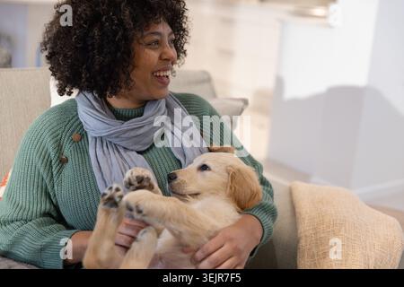 Afroamerikanerin sitzt zu Hause auf dem Sofa und hält goldene Retriever-Welpen in grünem Pullover. Haustier, Hund, gemütlich, Wärme, lockig, kni Stockfoto