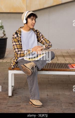 Asiatischer Teenager, der auf der Campus-Bank sitzt, Mütze und Headset trägt, Notebooks und Telefon hält. Holz, Lattenrost, Sitz, Outdo Stockfoto