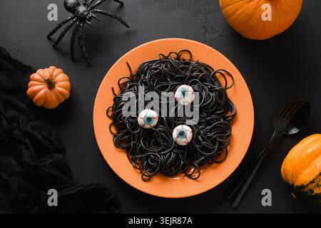 Halloween lustige italienische schwarze Pasta Tintenfisch Tinte dekoriert Horror Augen in orange Platte auf schwarzem Hintergrund. Horroressen für festliche Partys. Blick aus der ganzen Welt Stockfoto