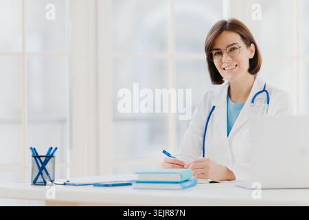Weibliche Ärztin schreibt Notizen am Schreibtisch in der medizinischen Praxis lächelnd KameraSchlüsselwörter: Doktor, Ärztin, Frau tun Stockfoto