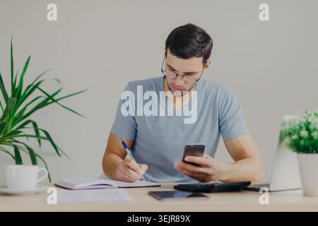 Junger Mann mit Smartphone und Notizen am Schreibtisch, der von zu Hause aus mit Laptop und Rechner arbeitet Stockfoto