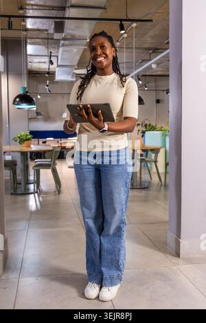 Afroamerikanerin, die im Coworking Space steht und ein Tablet hält, lächelt in Jeans und Smartwatch. Zusammenarbeit, Büro, Open-Air Stockfoto