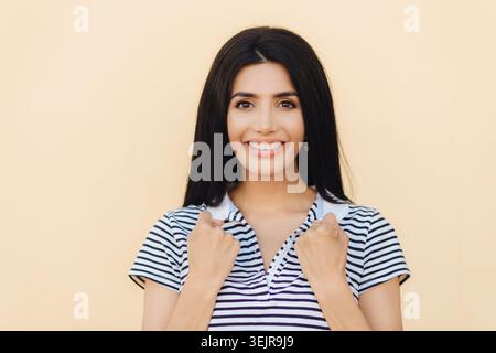 Lächelnde junge Frau in gestreiftem Hemd posiert auf beigefarbenem Studiohintergrund für lässige Mode- und Schönheitswerbung Stockfoto