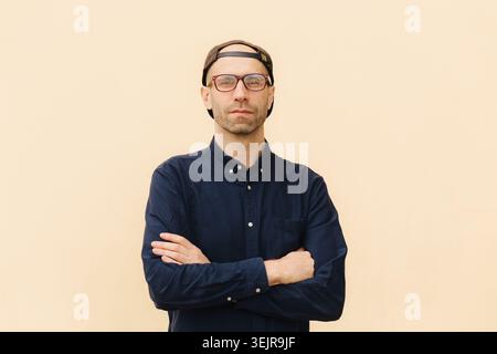 Ernster junger Mann mit gekreuzten Armen, Brille und Mütze auf beigefarbenem Hintergrund Stockfoto