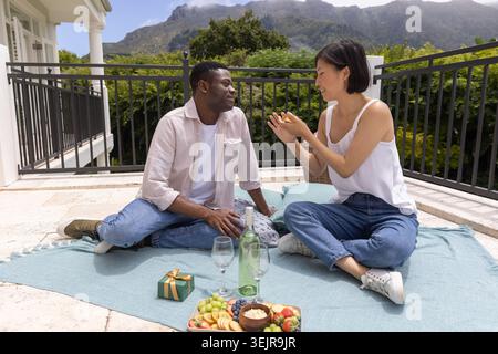 Verschiedene Paare sitzen auf hellblauer Picknickdecke auf der Terrasse und teilen sich Wein und Obstteller. Paar, Romantik, Balkon, draußen, Stockfoto