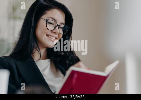 Junge Frau mit Brille, die zu Hause Buch liest und lächelt Stockfoto