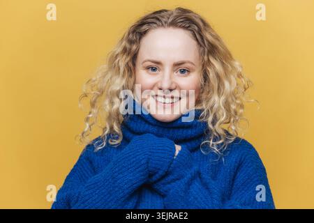 Lächelnde junge Frau in blauem Pullover und Schal isoliert auf gelbem Hintergrund für Wintermodeporträt Stockfoto