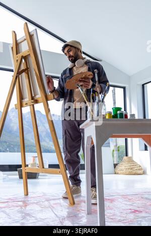 Mann aus den 30er Jahren malte an der Staffelei im Studio, trug Beanie und Kopfhörer, hielt Palette und Pinsel. Sonnenlicht, Maler, Atelier, Mount Stockfoto