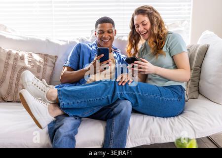 Verschiedene Paare sitzen auf hellem Sofa im hellen Wohnzimmer, halten Smartphones und lächeln. Duo, zu Hause, sonnenbeleuchtet, lässig, Stockfoto