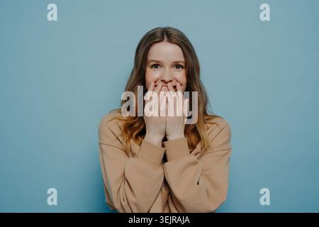 Überraschte junge Frau, die den Mund mit Händen auf blauem Hintergrund bedeckt Stockfoto