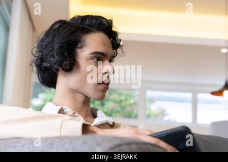Mann, der auf einem grauen Sofa im modernen Wohnzimmer sitzt und ein schwarzes Smartphone hält, während er eine beige Jacke trägt. Porträt, Seitenansicht, Telefon, Innenraum, Zuhause, Pendan Stockfoto