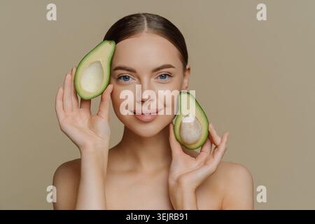 Junge Frau, die frische Avocadohälften nahe Gesicht auf Beige Hintergrund für natürliche Schönheit und gesunde Haut hält Stockfoto