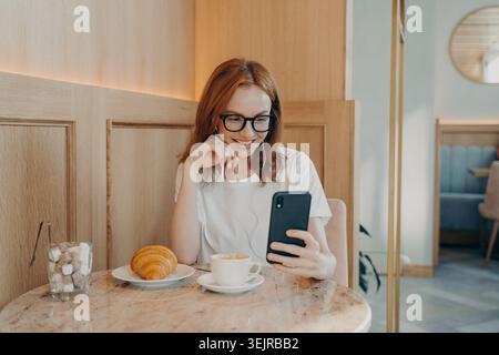 Junge Frau mit Smartphone im Café, die am Tisch sitzt und Croissant lächelt Stockfoto