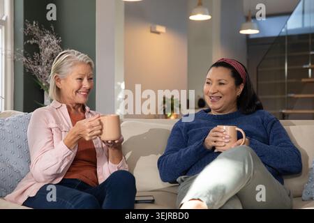 Ältere Freundinnen schlürfen an Keramikbechern und plaudern mit dem Smartphone auf dem Sofa im Wohnzimmer. Alter, Begleitung, Konversation Stockfoto