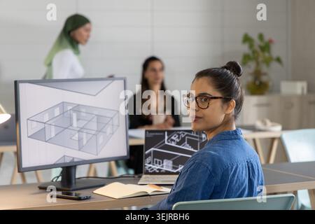 Asiatische Frau in Denim-Hemd, die sich am offenen Schreibtisch zur Kamera dreht und den 3D-Grundriss auf dem Monitor überprüft. Büro, Zusammenarbeit, Arch Stockfoto