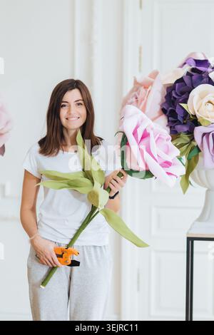 Junge Frau, die riesige Papierblumendekorationen im hellen Studio-Workshop-Innenraum hält Stockfoto